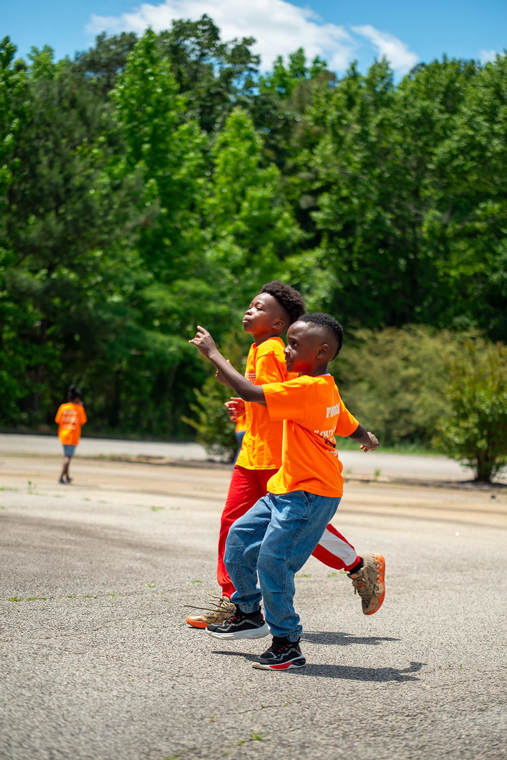 Two children in orange shirts walk and play on a sunny playground area with trees in background.